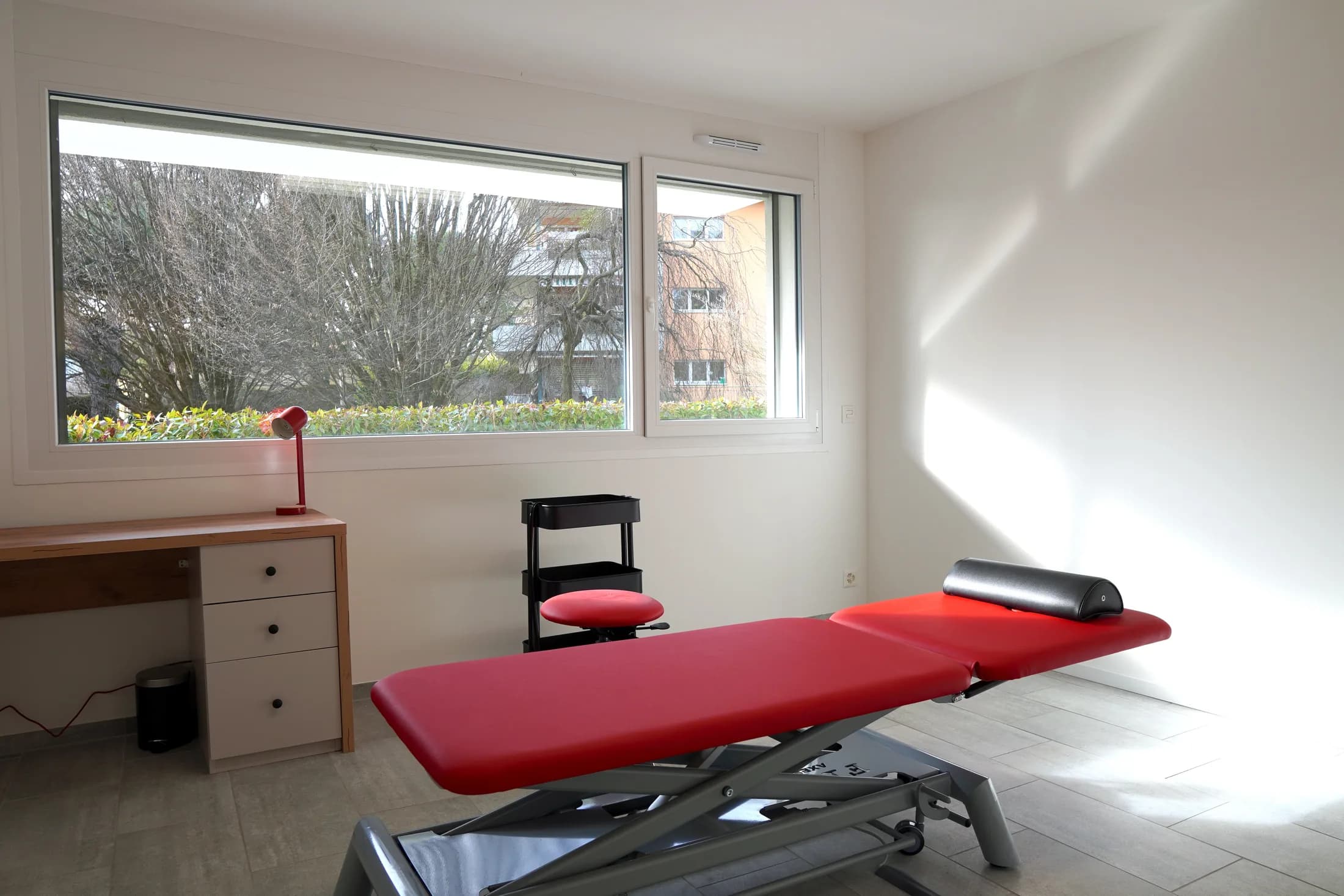Salle de traitement lumineuse, table de soins rouge et bureau devant la fenêtre