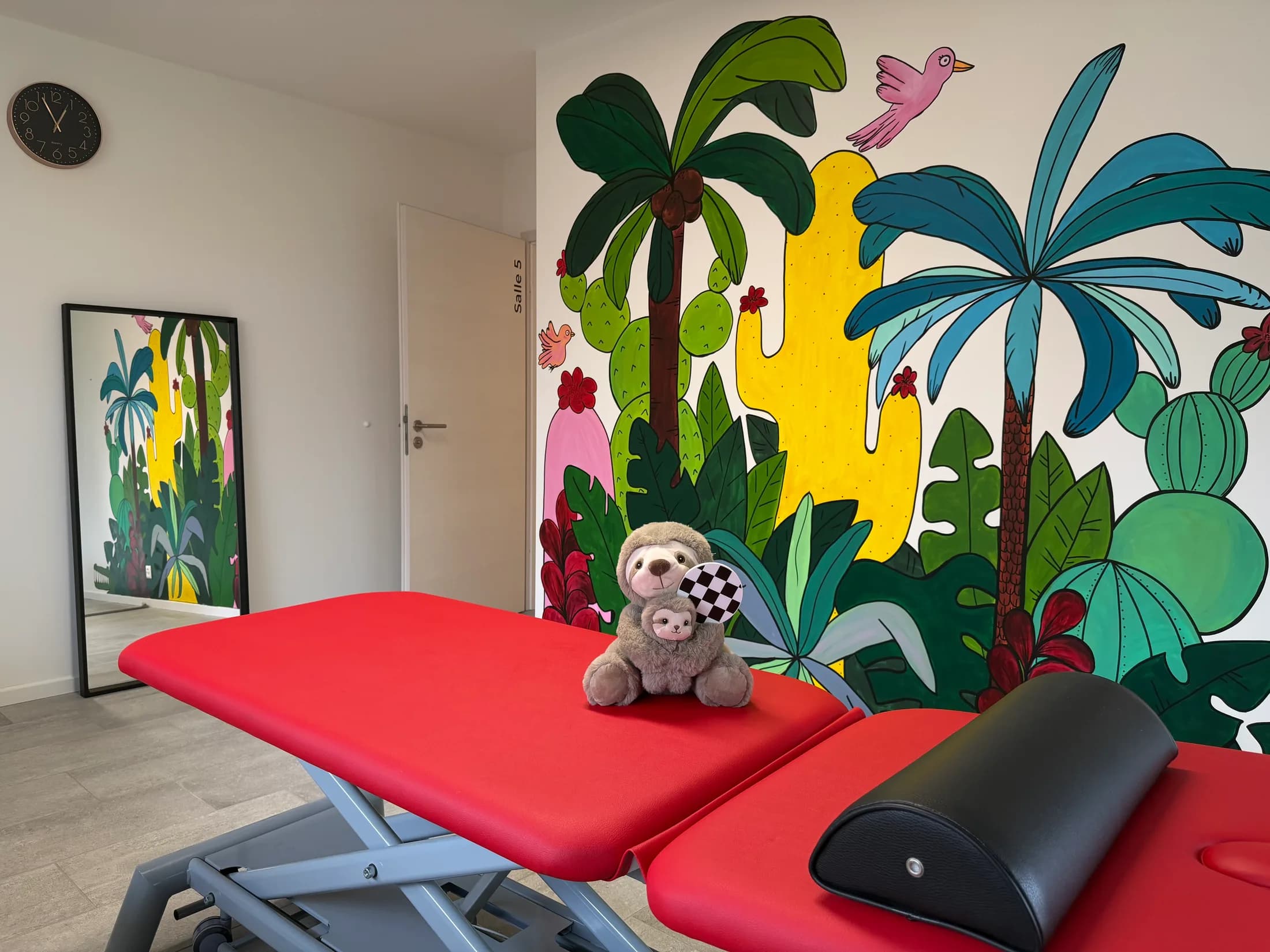 Salle de physiothérapie pédiatrique, table de soins rouge devant une fresque tropicale